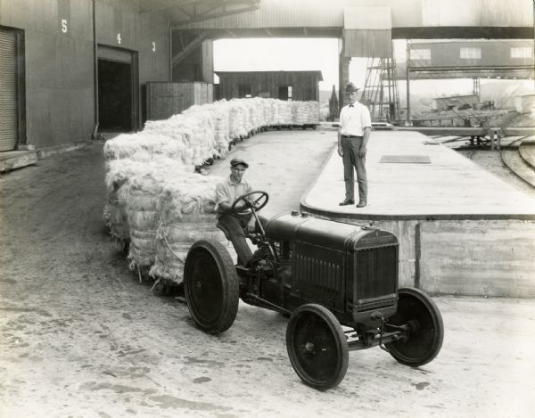 A man operating a McCormick-Deering 10-20 industrial tractor to haul a long train of carts holding bundles of fiber, possibly for burlap. Another man is looking on from a raised platform.