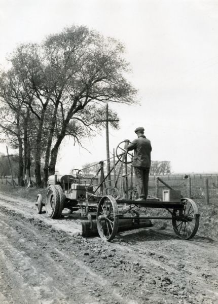 A man stands while operating a McCormick-Deering 10-20 industrial tractor and an International duplex grader to smooth a rural dirt road. The original caption reads: "Owned by Lee County, Illinois and operated by Fred Barnhizer, Polo, Ill. This outfit is covering a 30 mile rectangle, 10 miles long and 5 miles wide. The 30 mile round trip is usually made in one day and the other side of the road is gone over the following day. The road is kept in A-1 shape."