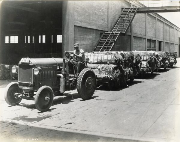 A man operating a McCormick-Deering 10-20 industrial tractor to haul carts loaded with what appear to be bundles of burlap fabric. There is an industrial building in the background, and a stairway leading to a walkway over the road.