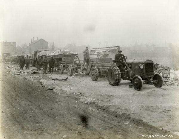 Men stand alongside a line of machinery and vehicles near a dirt road, including a McCormick-Deering 10-20 industrial tractor, at what appears to be a construction site. There are buildings in the background.