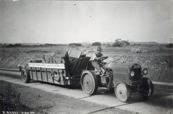 A man uses a McCormick-Deering 10-20 industrial tractor to haul a wagon full of wooden boards along a paved road. The sign on the wagon reads: "San Diego Consolidated Gas & Electric Co."