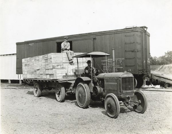 Two men from the Lexington Hardware Corporation use a McCormick-Deering industrial 10-20 tractor to haul a wagon loaded with boxes of metal shingles. A train car and railroad tracks are in the background.