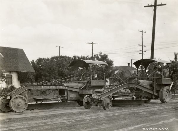 Men use McCormick-Deering 10-20 industrial tractors with Russell graders to smooth a dirt road. There are houses and power lines in the background.