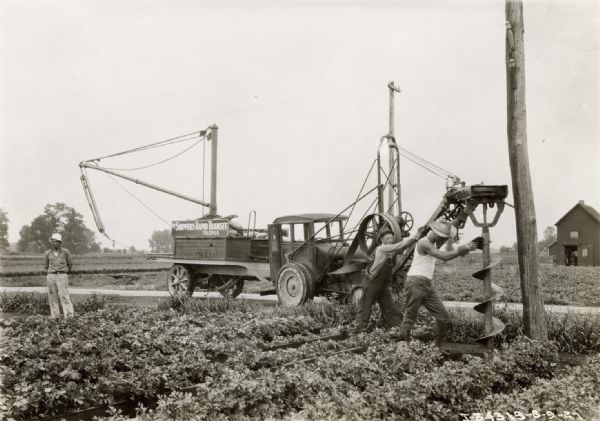 Men use a modified McCormick-Deering 10-20 industrial tractor with an attached trailor and post hole digging rig auger to install a telephone or electric pole alongside a road.