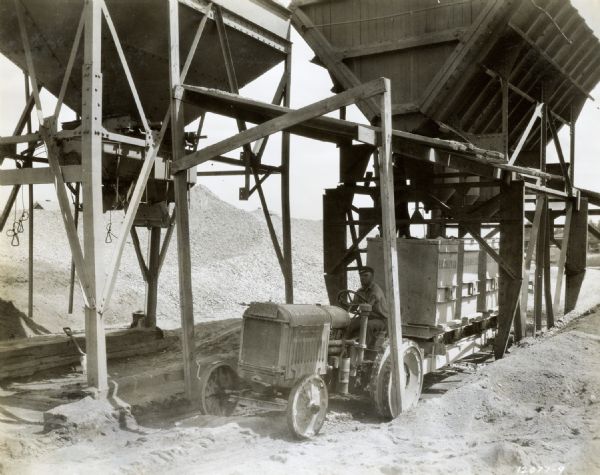 An employee of the R.D. Baker Company uses a McCormick-Deering 10-20 industrial tractor to load batch boxes with sand, cement, and gravel from large overhead bins.
