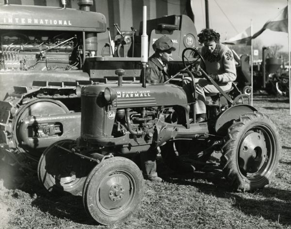 A woman is sitting behind the wheel of a McCormick-Deering Farmall Cub tractor for a demonstration at International Harvester's Hinsdale experimental farm. A man smoking a cigarette is standing beside her.