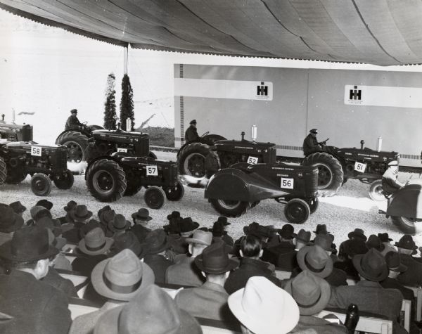 An audience watches as men demonstrate a variety of tractors beneath a tent on International Harvester's Hinsdale experimental farm. Tractors include the W-9, WD-9, O-6 (orchard), WD-6, and the OS-4.