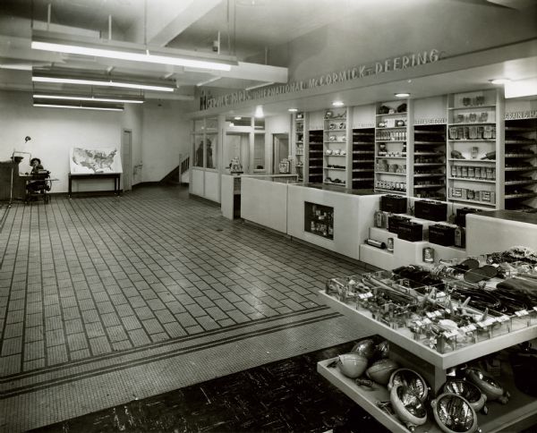 Interior view of International Harvester's Education and Training Center. Machinery parts and equipment are on display along the wall of a service and parts area. The room looks like a model of a company dealership. A woman is sitting near the back of the room near a map of the United States.