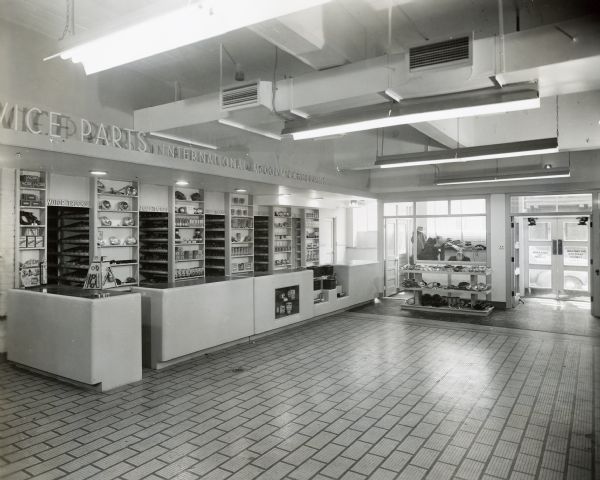 Interior view towards entrance of the service and parts area of International Harvester's Education and Training Center. Two men in an office are in the background.