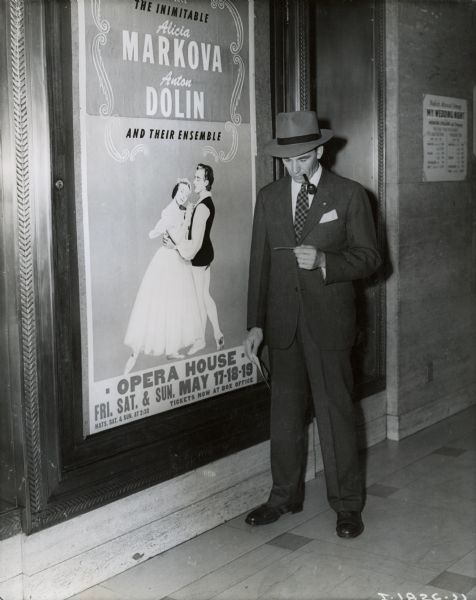 A man stands near an opera house sign, smoking a pipe and looking at his theater ticket. A poster advertises a show by Alicia Markova and Anton Dolin.