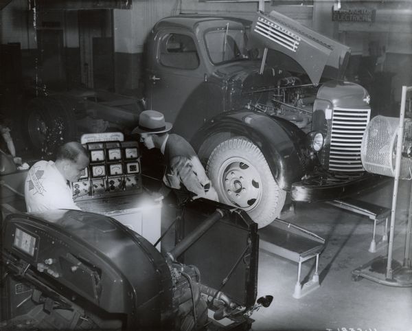 A man in a suit and hat looks on as another man in an International Harvester shirt demonstrates a Sun Motor Tester at International Harvester's Education and Testing Center.