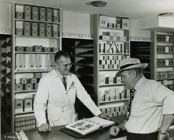 A man in an International work coat is standing behind a counter and showing another man a binder with photographs of machinery parts. A storage area of machinery parts and boxes is behind the men. The original caption reads: "Paul Dean & Harold Carr, IH personnel."