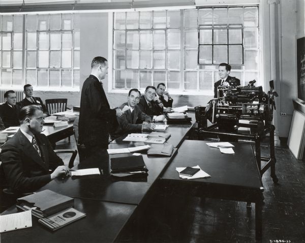 A man stands behind a table and addresses a teacher in a classroom at International Harvester's Education and Training Center. The teacher sits beside an engine near a window, and other students sit behind tables in the classroom.