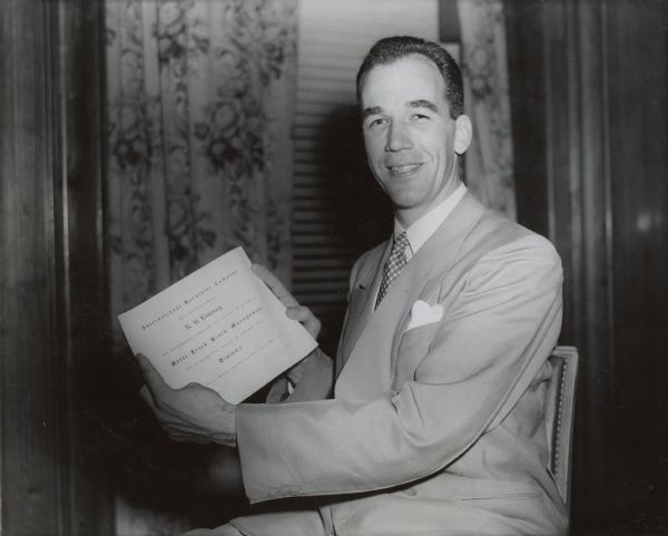 A man wearing a suit holds up a diploma in Motor Truck Block Management from International Harvester's Education and Training Center. The diploma reads: "International Harvester Company; This Certifies that R.S. Livesay has successfully completed the course of study in Motor Truck Block Management and in recognition thereof is awarded this Diploma."