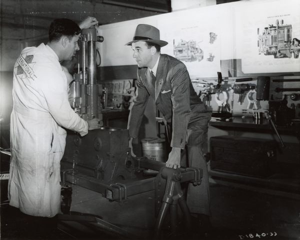 A man wearing an International work coat demonstrates an engine for a man wearing a suit and hat. The men are at International Harvester's Education and Training Center. Posters of engines and engine parts hang on the walls in the background.
