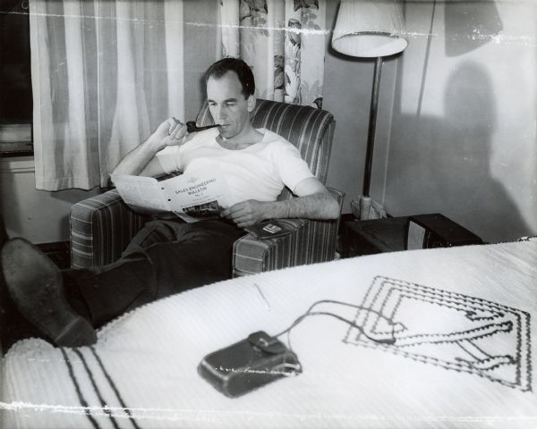 A man sitting in an armchair with his feet propped on a bed, is reading an International sales-engineering bulletin and smoking a pipe.