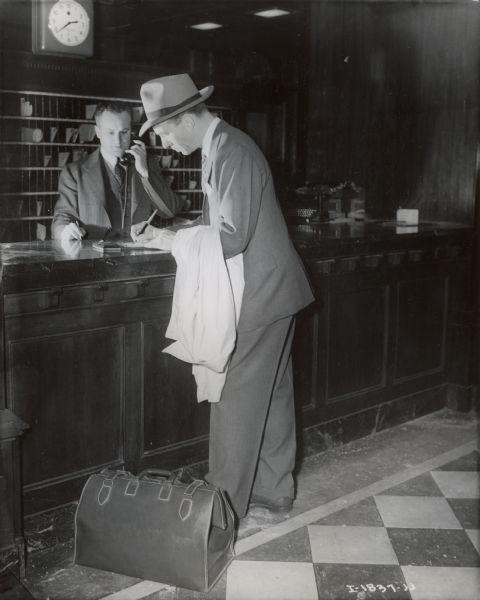 A man wearing a suit and hat is checking in and signing a piece of paper at a hotel registry desk. A suitcase is sitting at his feet and a hotel employee is using a telephone behind the desk.