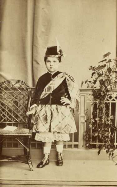 Full-length studio portrait of a boy standing and resting his hand on a chair, possibly Stanley McCormick (1874-1947). He is posing in what appears to be a Scottish outfit, including a hat, and a jacket with a sash.