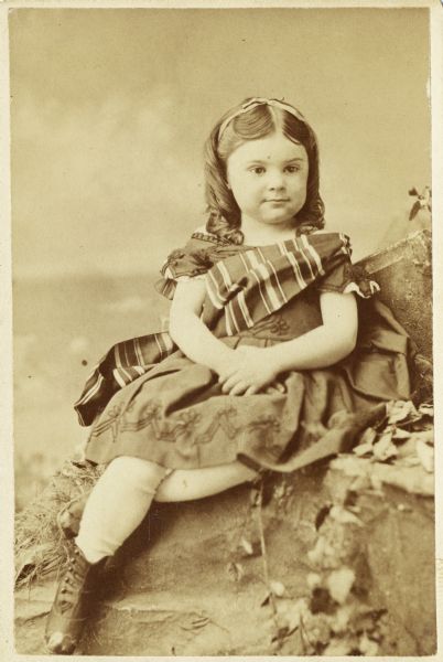 Studio portrait of Anita McCormick Blaine (1866-1954) seated on a prop rock outcropping. She is wearing a dress with a sash over her shoulder, with leggings and boots.