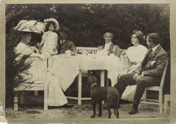 A group of men, women, and children, including Nettie Fowler McCormick sitting at a table outdoors. A dog is standing in the foreground, and tea cups and containers of food are on the table.