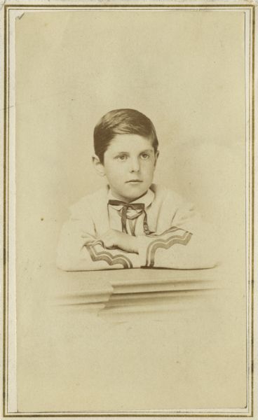 Studio portrait of a boy, possibly Cyrus McCormick, Jr., with his arms crossed on a table. He is wearing a decorated jacket and necktie.