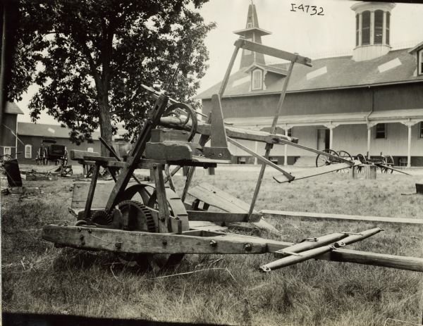 Side view of a McCormick reaper resting on grass near a large barn and other farm buildings. The original caption reads: "Invented 1845, patented 1847. The improvement embodied in this machine is a seat for carrying the raker, and which enabled him, while riding, to rake the grain from the platform, depositing it in gavels on the ground."