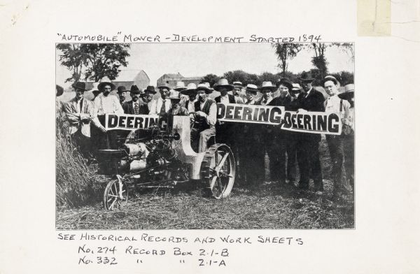 A group of men outdoors are standing behind a Deering Auto-Mower holding Deering signs. The mower was an experimental gas powered mowing machine. The text above the photograph reads: "'Automobile' Mower - Development Started 1894."