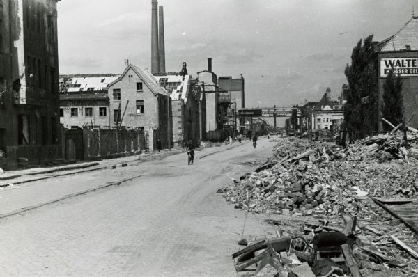 A man rides a bicycle down a road at International Harvester's Neuss Works, surrounded on either side by rubble and buildings. The factory was heavily damaged during World War II. The original photograph caption reads: "Taken at the entrance of Industriestrasse."
