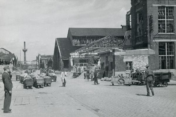 Men are walking down the center road at International Harvester's Neuss Works. They are surrounded on either side by buildings, possibly war-damaged.