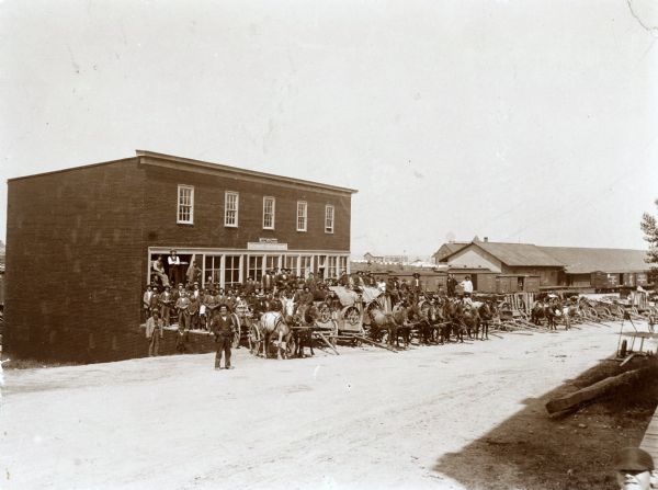 A group of men with horse-drawn wagons gathering in front of a McCormick Harvesting Machine Company dealership. The wagons appear to be loaded with farm implements. The gathering may be part of a "McCormick Day" promotion, in which customers gather on an appointed to day to pick up the machines they've ordered. Railroad cars are in the background.