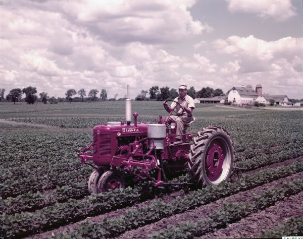 View towards a man using a McCormick Farmall Super C tractor with a C-254 cultivator in a soybean field. Farm buildings are in the background. The original caption reads: "C-254 cultivator with tooling equipment on Farmall Super C with No.310 fertilizer unit.  Used in soybeans."