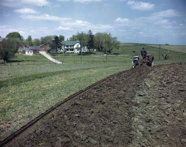 View across field towards a man using a Farmall H tractor with a 2-furrow trailing plow to work in a field.  A farmhouse and several outbuildings are in the background. The original caption reads: "Farmall H tractor with a No.8 2-furrow plow and a No.46 plowsole fertilizer attachment plowing on the contour."