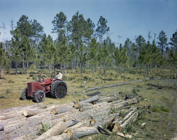 Slightly elevated view of a man using a specially outfitted International tractor to work among felled trees in a wooded area. The original caption reads: "Two specially fitted tractors, and ID-9 and an I-4 are shown at work on a pulpwood project near Nahunta, Ga. for the Brunswick Pulp and Paper Co."