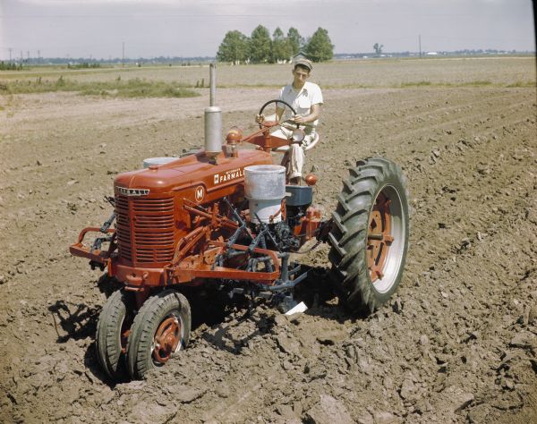 Slightly elevated view of a man using a McCormick Farmall M tractor with a forward mounted cotton planter to work in a field. The original caption reads: "HM-277 blackland planter equipped with Type C Duplex hopper and fertilizer. Used on a Farmall M tractor."