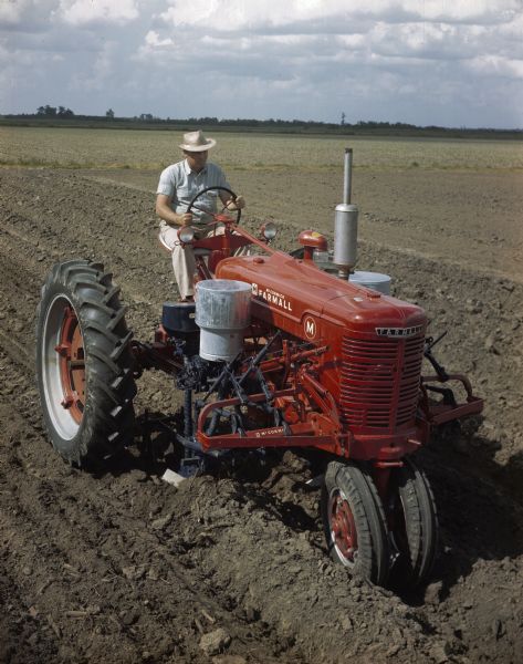 Slightly elevated view of a man using a McCormick Farmall M tractor with a forward-mounted cotton planter attachment to work in a field. The original caption reads: "HM-277 blackland planter equipped with Type-C Duplex hopper and fertilizer. Used on a Farmall M tractor."