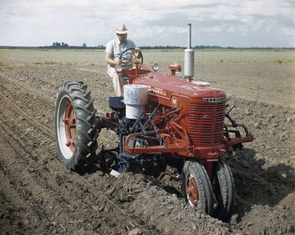 Slightly elevated view of a man using a McCormick Farmall M tractor with a forward-mounted cotton planter to work in a field. The original caption reads: "HM-277 blackland planter equipped with Type C Duplex hopper and fertilizer. Used on a Farmall M tractor."