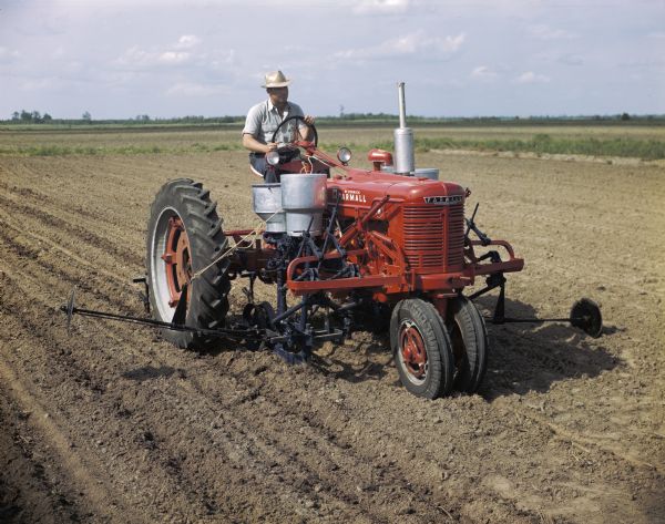 View towards a man using a McCormick Farmall H tractor with a forward-mounted cotton planter to work in a field. The original caption reads: "'Flat' planting with HM-278 runner type planter equipped with single-seed cotton hopper and fertilizer unit with runner applicator."
