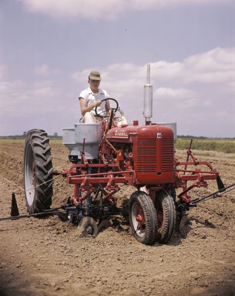 View towards a man using a Farmall C tractor with a forward-mounted cotton planter to work in a field. The original caption reads: "'Furrow' planting with C-278 runner type planter equipped with special disk furrowing attachment, single-seed cotton hopper and fertilizer unit with deep applicator."