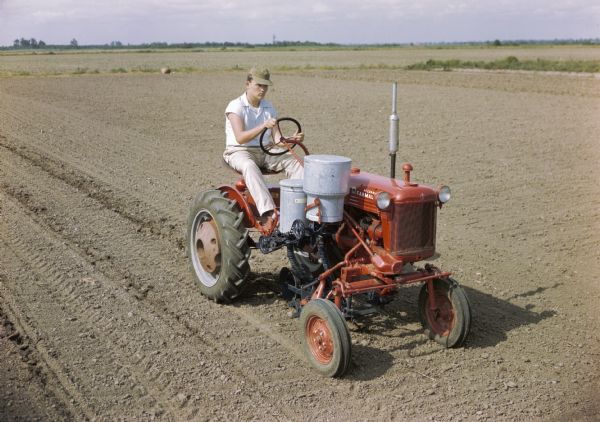 Slightly elevated view of a man using a Farmall Cub with a forward-mounted cotton planter attachment to work in a field. The original caption reads: "Planting cotton on the 'flat' with Cub-172 runner type planter equipped with single-seed cotton hopper, regular ground tools, and fertilizer unit with deep applicator."