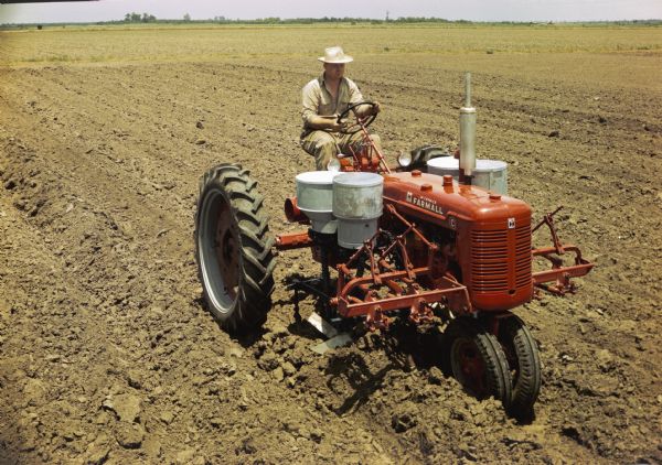 Slightly elevated view of a man using a Farmall C tractor with a cotton planter attachment to plant cotton in a field. The original caption reads: "C-277 blackland planter equipped with single-seed cotton hopper and fertilizer unit."