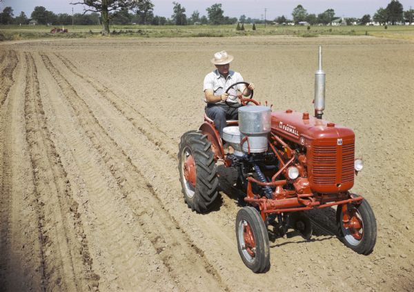 Slightly elevated view of a man using a Farmall Super A tractor with a cotton planter attachment to plant in a field. The original caption reads: "'Bed' planting with A-278 runner-type planter equipped with disk bedding attachment, reverse-feed cotton hopper and fertilizer unit."