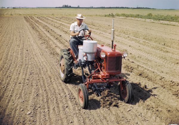 Slightly elevated view of a man using a McCormick Farmall Cub tractor with a forward-mounted cotton planter to work in a field. The original caption reads: "'Furrow planting' cotton with Cub-172 runner-type planter equipped with single-seed cotton hopper, fertilizer unit, and regular ground unit with special shovel furrowing attachment."