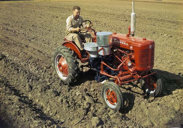 Color photograph of a man using a McCormick Farmall Super A tractor with a forward mounted cotton planter to work in a field. The original caption with the color photograph reads: "'Blackland' planting with A-277 planter equipped with reverse-feed cotton hopper and fertilizer unit."