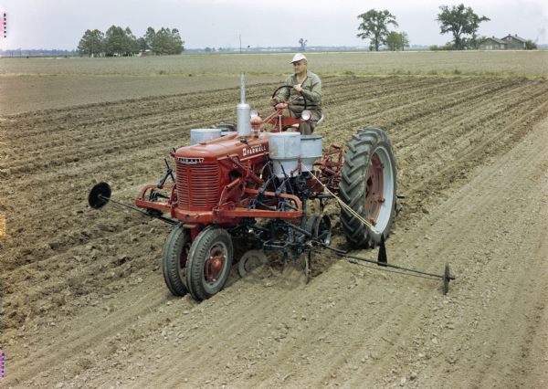Slightly elevated view of a man using a McCormick Farmall H tractor with a HM-278 cotton planter to work in a field. The original caption reads: "'Bed' planting with HM-278 runner type planter equipped with disk bedding attachment, single-seed cotton hopper and fertilizer units with runner applicators."