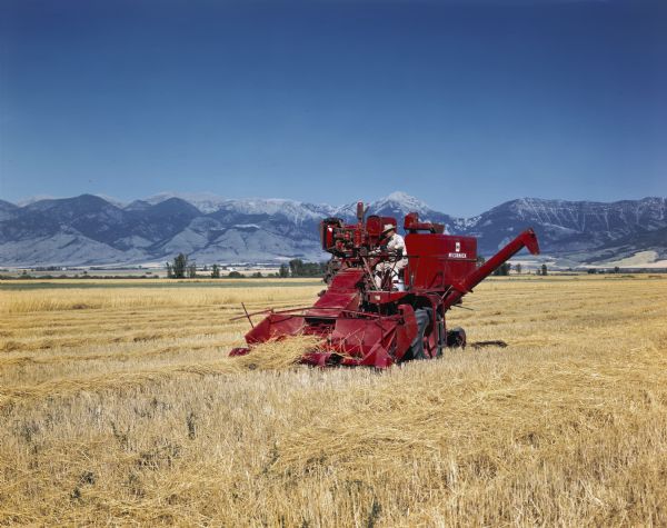 Side view of a man using an experimental No.127 SP harvester-thresher (combine) in a field.  Mountains are in the far background. The original caption reads: "These pictures were taken of the experimental self-propelled 125-SPX and the pull type 122-C combines and are to be used only with the permission of the sales and engineering departments."