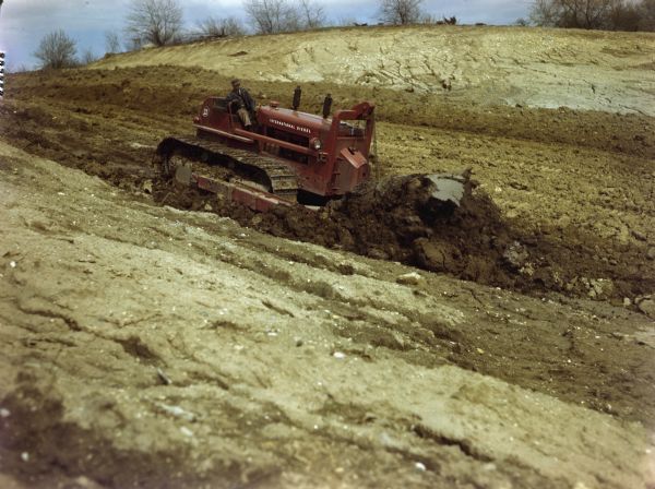 View towards a man using an International Diesel TD-24 crawler tractor (TracTracTor) with a BE bullgrader to work in a field. The original caption reads: "Photographed at Fin and Feather Club."
