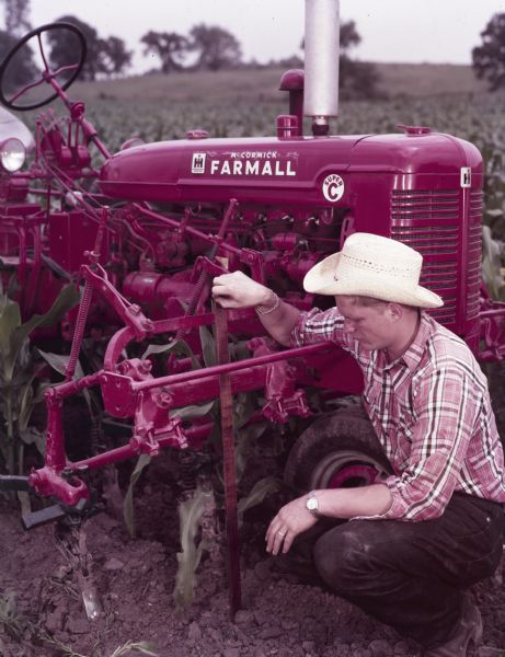 View of a man crouching in front of a McCormick Farmall Super C tractor with a C-254 cultivator to measure a stalk of corn in a field.