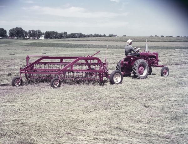 View towards a man using a McCormick Farmall Super A tractor with a No.5-LW side delivery rake to work in a field. The original caption reads, "Windrowing hay with No.5-LW Side Delivery Rake."