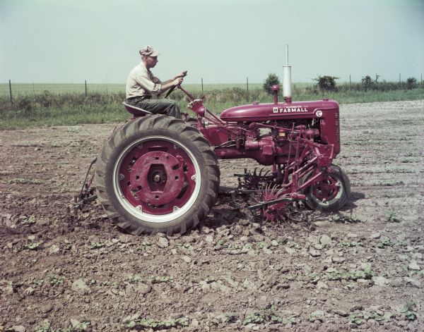 View towards a man using a McCormick Farmall Super C tractor with a C-254 cultivator to work in a field. The original caption reads: "C-254 two-row cultivator with No.10 rotary weeder on Super C. First cultivation of cotton at Hinsdale, breaking hard crust in the rows."