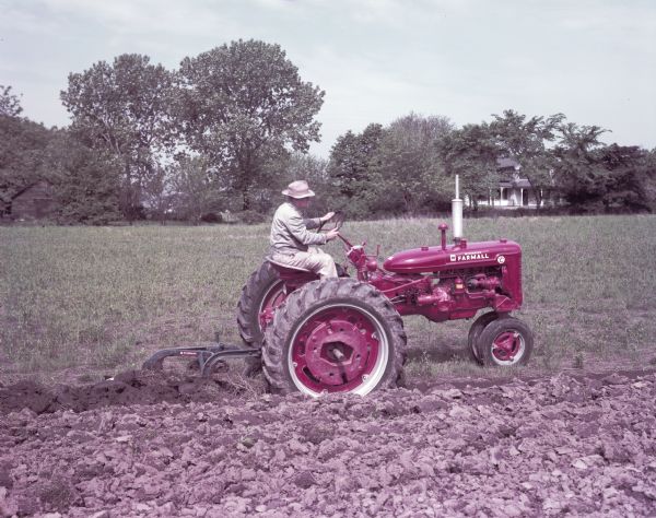 View towards a man using a McCormick Farmall Super C tractor with a C-295A plow. The original caption reads: "Farmall Super C and McCormick C-295A plow with 14-inch Plow Chief bottoms."